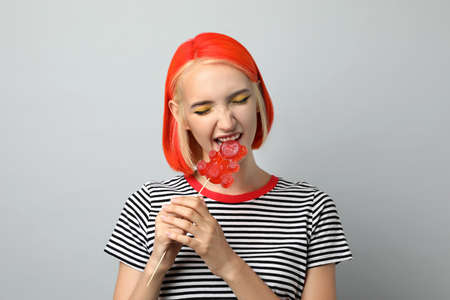 Beautiful young woman with bright dyed hair eating bear shaped candy on light grey background