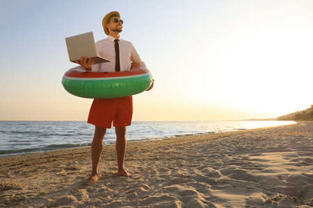 Happy man with inflatable ring and laptop on beach, space for text. Business tripの写真素材