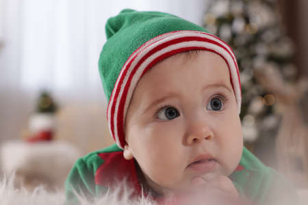 Baby wearing cute elf costume in room decorated for Christmas