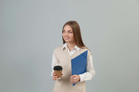 Teenage student with folder and paper cup of coffee on grey background