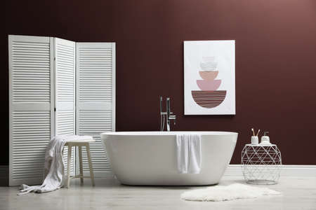 Modern ceramic bathtub and furniture near burgundy wall in roomの写真素材