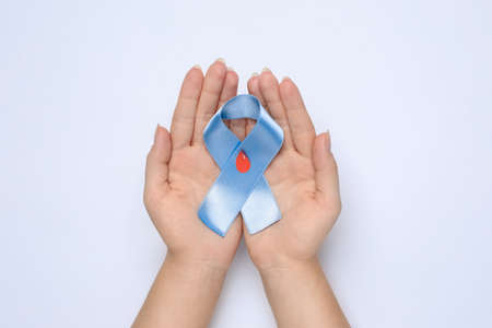 Woman holding light blue ribbon with drop of blood on white background, top view. Diabetes awareness