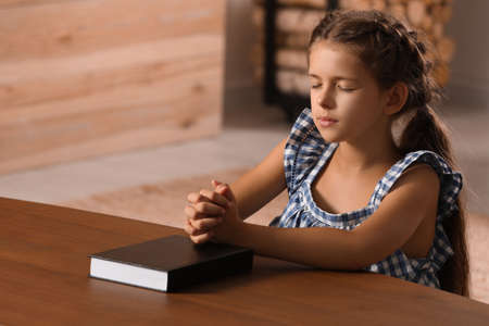 Cute little girl praying over Bible at table in roomの写真素材