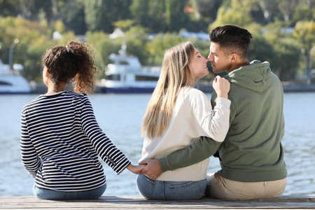 Man holding hands with another woman behind his girlfriend's back on pier near river. Love triangleの写真素材