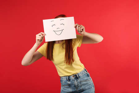 Woman hiding behind sheet of paper with happy face on red backgroundの写真素材