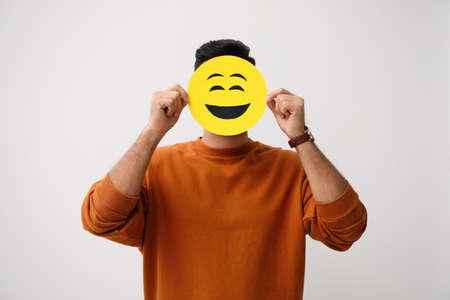 Man hiding emotions using card with drawn smiling face on white backgroundの写真素材
