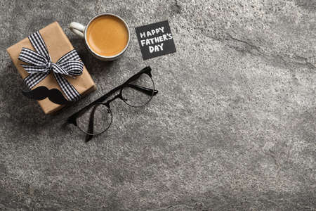 Card with phrase HAPPY FATHER'S DAY, gift box, eyeglasses and cup of aromatic coffee on gray background, flat lay. Space for textの写真素材