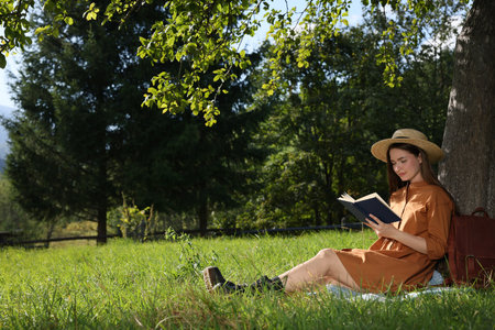 Young woman reading book under tree on meadow near forestの写真素材