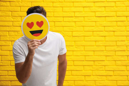 Man hiding emotions using card with drawn smiling face near yellow brick wall. Space for textの写真素材