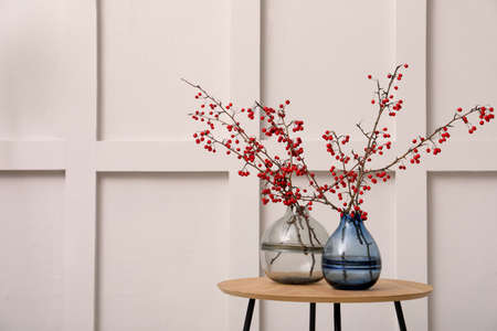 Hawthorn branches with red berries in vases on wooden table near light wall indoors, space for textの写真素材