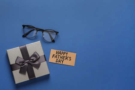 Card with phrase HAPPY FATHER'S DAY, eyeglasses and gift box on blue background, flat lay. Space for...