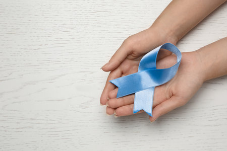 Woman holding light blue awareness ribbon at white wooden table, top view. Space for textの写真素材