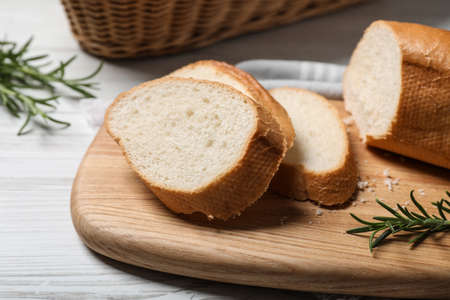 Cut tasty baguette with rosemary on white wooden table, closeup