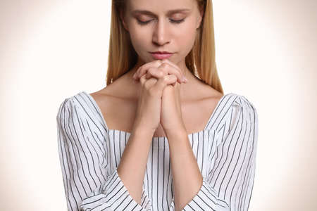 Religious young woman with clasped hands praying against light background, closeupの写真素材