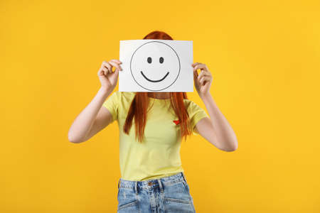 Woman hiding behind sheet of paper with happy face on yellow backgroundの写真素材
