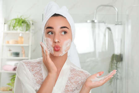 Beautiful young woman applying cleansing foam onto face in bathroom. Skin care cosmeticの写真素材