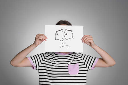 Woman hiding behind sheet of paper with sad face on gray backgroundの写真素材