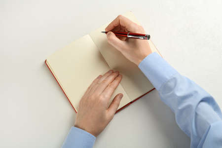 Man writing with pen in notebook at white table, top view