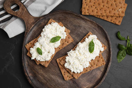 Crispy crackers with cottage cheese and basil on black table, flat lay