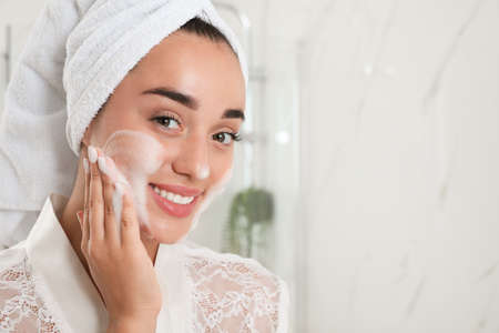 Beautiful young woman applying cleansing foam onto face in bathroom, space for text. Skin care...