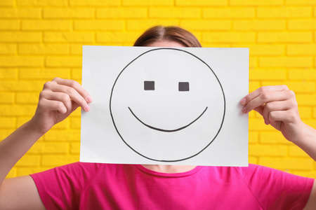 Woman hiding behind sheet of paper with happy face against yellow brick wallの写真素材