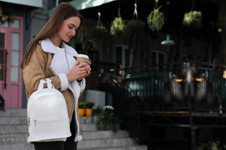 Young woman with stylish white backpack and cup of drink on city street, space for text