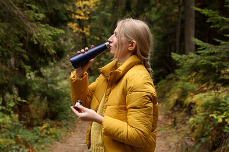 Young woman drinking hot beverage from thermo bottle in autumn forest