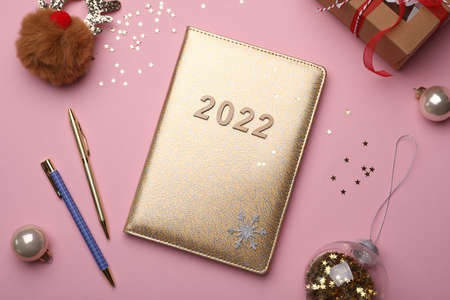 Golden planner with number 2022, stationery and festive decor on pink background, flat lay. New Year...