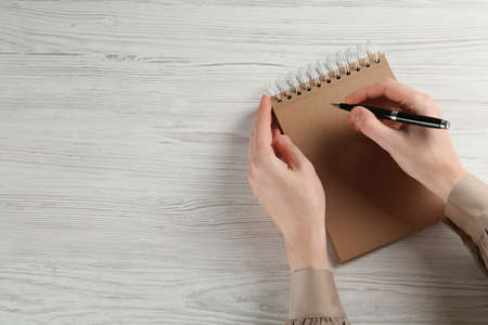 Woman writing with pen in notebook at white wooden table, top view. Space for text
