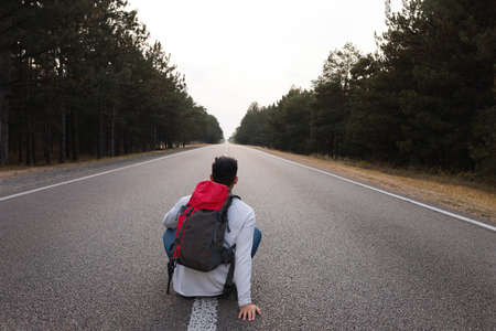 Man with backpack sitting on road near forest, back viewの写真素材