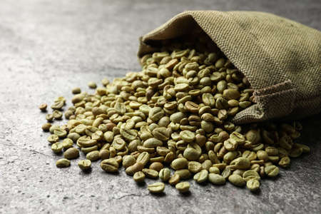 Overturned sackcloth bag with green coffee beans on gray table, closeup