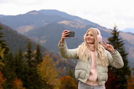 Young woman in warm earmuffs taking selfie on autumn day