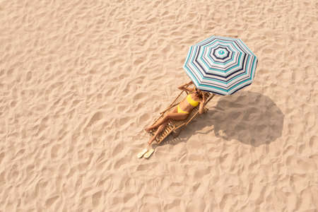 Woman resting in sunbed under striped beach umbrella at sandy coast, space for textの写真素材
