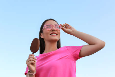 Beautiful young woman holding ice cream glazed in chocolate against blue sky, low angle view