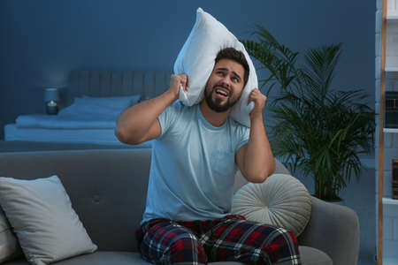 Unhappy young man covering ears with pillow in living room at night. Noisy neighbors