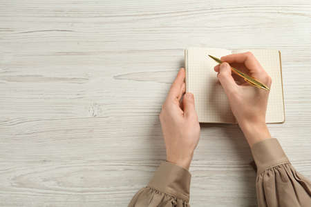 Woman writing with pen in notebook at white wooden table, top view. Space for text
