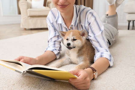 Young woman reading book with cute dog on floor in living room, closeupの写真素材