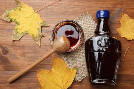 Bottle of tasty maple syrup, bowl and dry leaves on wooden table, flat layの写真素材