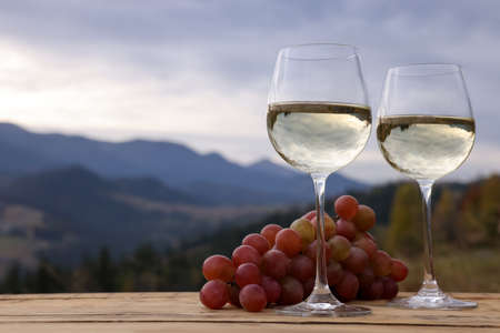 Glasses of tasty wine and grapes on wooden table against mountain landscape. Space for textの写真素材