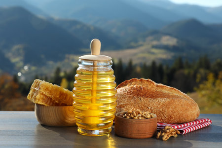 Fresh aromatic honey, combs, bread and nuts on gray wooden table against mountain landscapeの写真素材