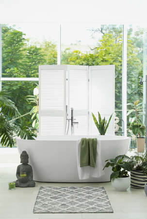 Stylish bathroom interior with modern tub, window and beautiful houseplants. Home design