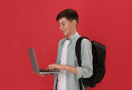 Teenage student with backpack and laptop on red background
