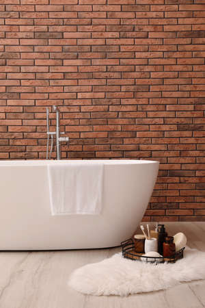 Modern ceramic bathtub and tray with toiletries near brick wall indoors