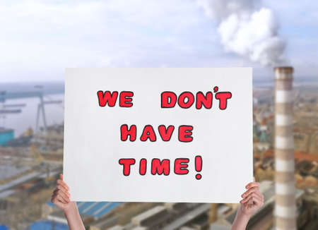 Protestor holding placard with text We Don't Have Time and blurred view of industrial factory on...