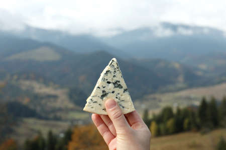 Woman holding piece of delicious cheese against mountain landscape, closeupの写真素材