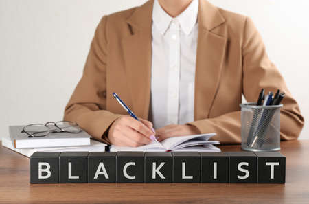 Black cubes with word Blacklist and woman working at wooden office desk, closeupの写真素材