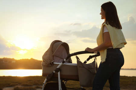 Happy mother with baby in stroller walking near river at sunsetの写真素材