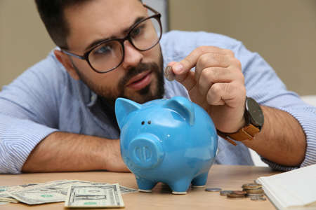 Young man putting coin into piggy bank at table indoors, focus on hand. Money savingsの写真素材