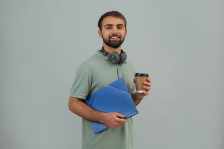 Student with headphones, folder and paper cup of coffee on gray background