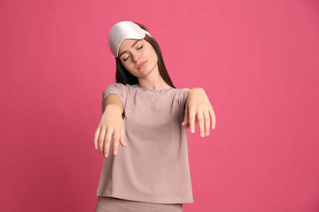 Young woman wearing pajamas and mask in sleepwalking state on pink background